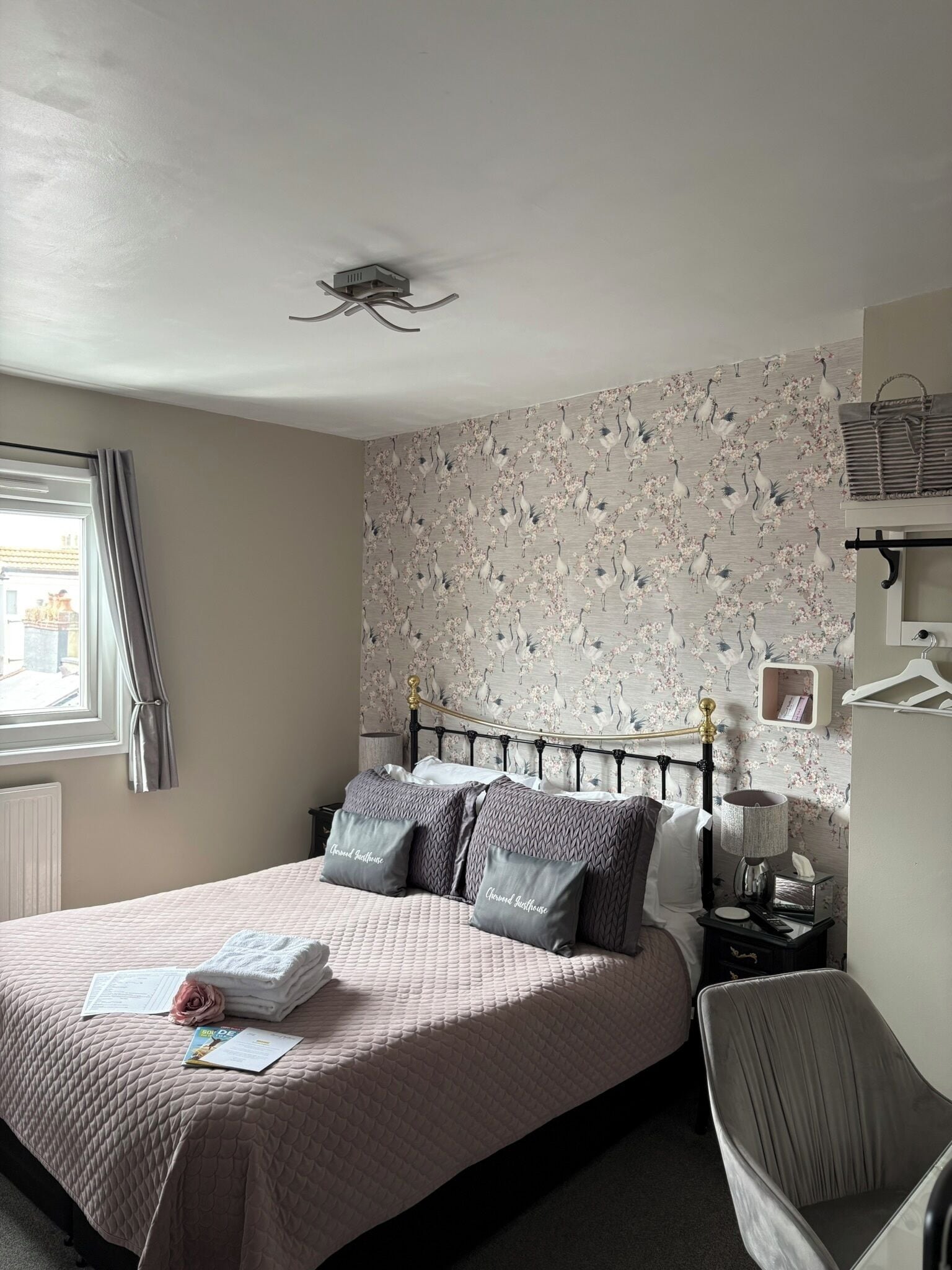 A neatly made bed with pillows, a bedside table with a lamp, a chair, and a wall with a floral pattern.