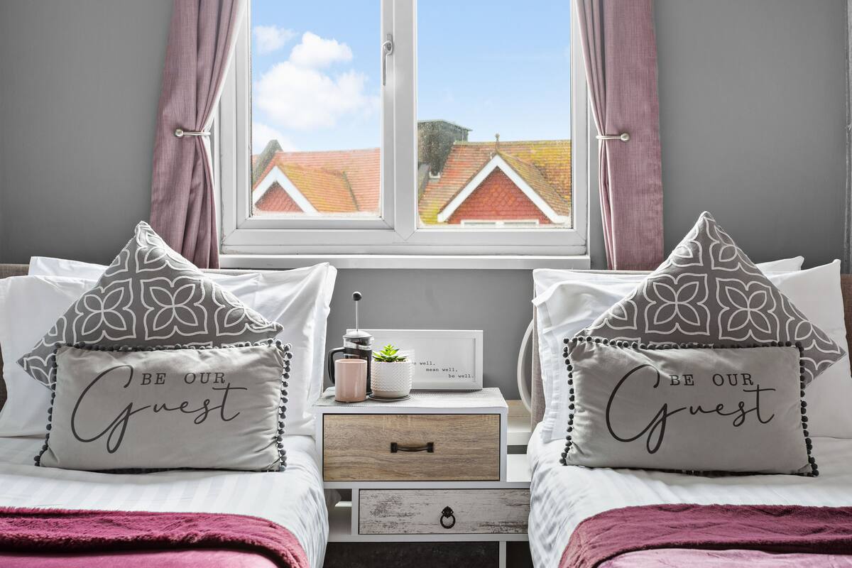 Une chambre avec deux lits, chacun agrémenté d’un coussin sur lequel est inscrit : « SOYEZ LES BIENVENUS ». Il y a une table de chevet avec une petite plante et un tableau encadré placé entre les deux lits.