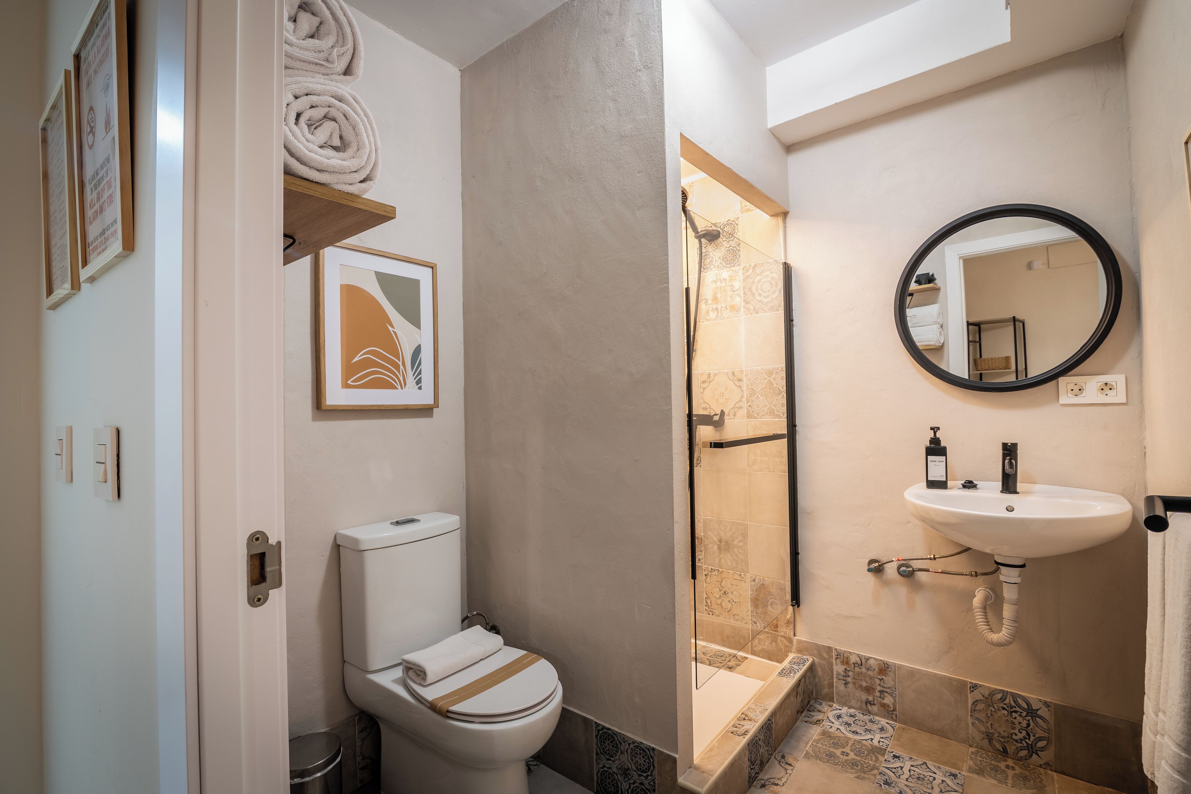 Un baño con inodoro, lavamanos y ducha. Hay toallas en un estante, un cuadro en la pared y un espejo sobre el lavamanos.