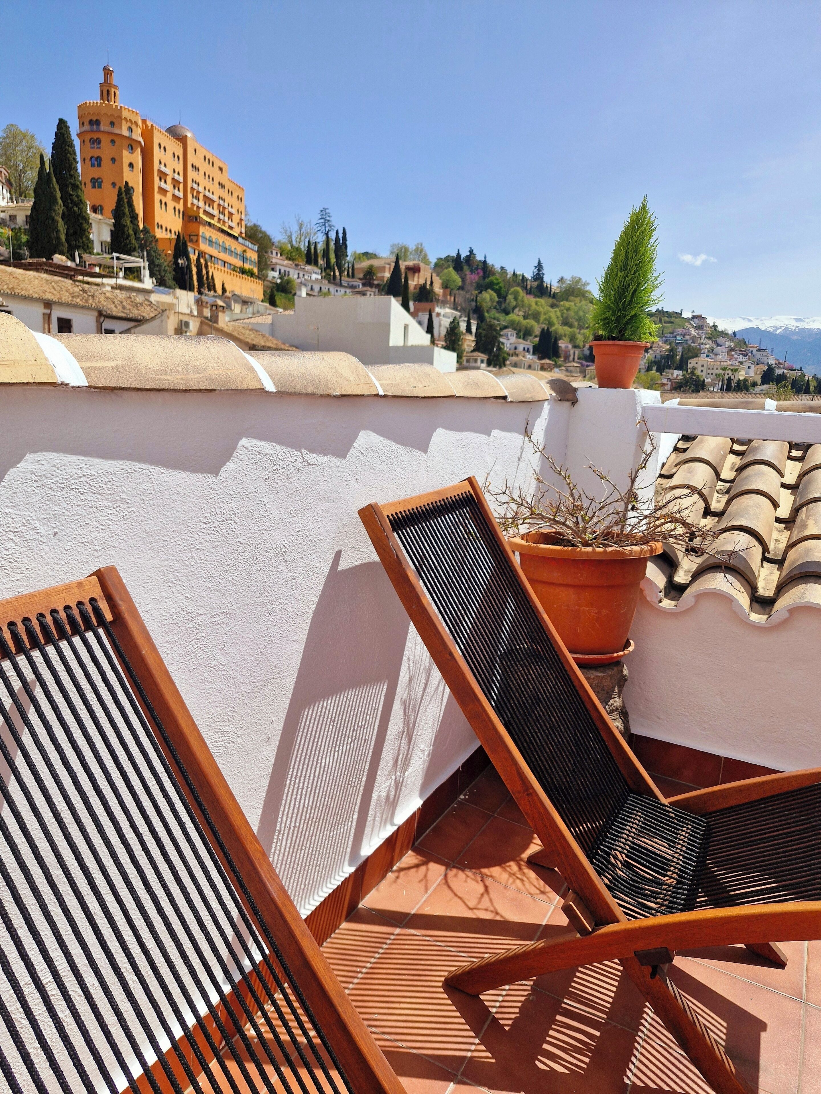 A rooftop terrace with two wooden lounge chairs, a potted plant, and a view of a historic building and mountains in the distance.