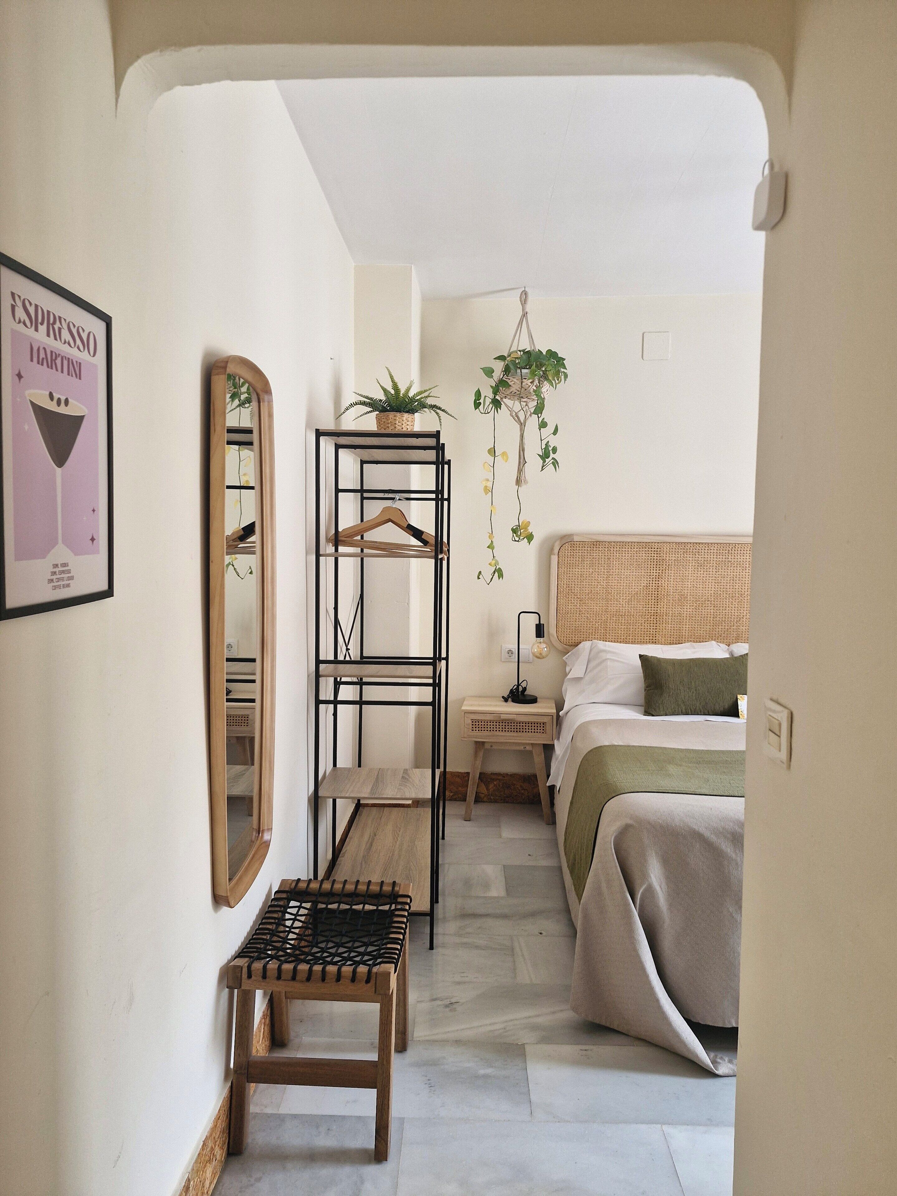 A bedroom with a bed, a bedside table, a hanging plant, a mirror, and a framed picture on the wall.