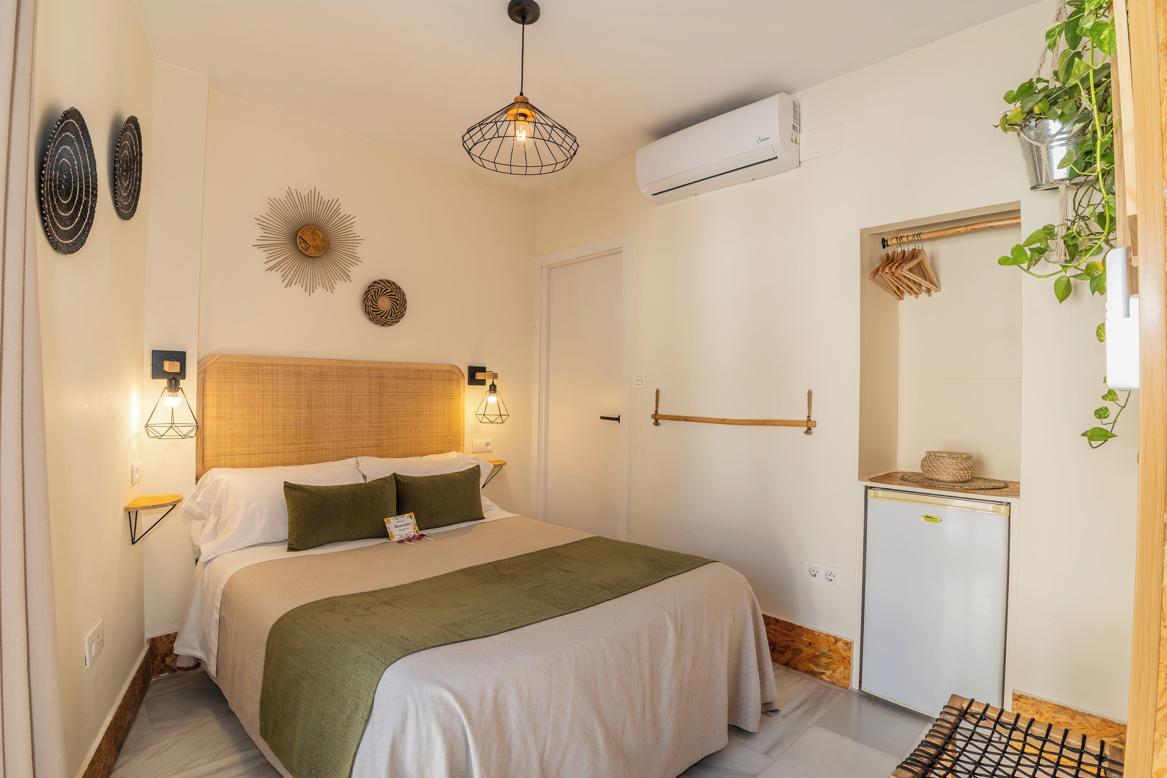 Un dormitorio con cama, lámparas de noche, aire acondicionado de pared, un refrigerador pequeño y una planta.