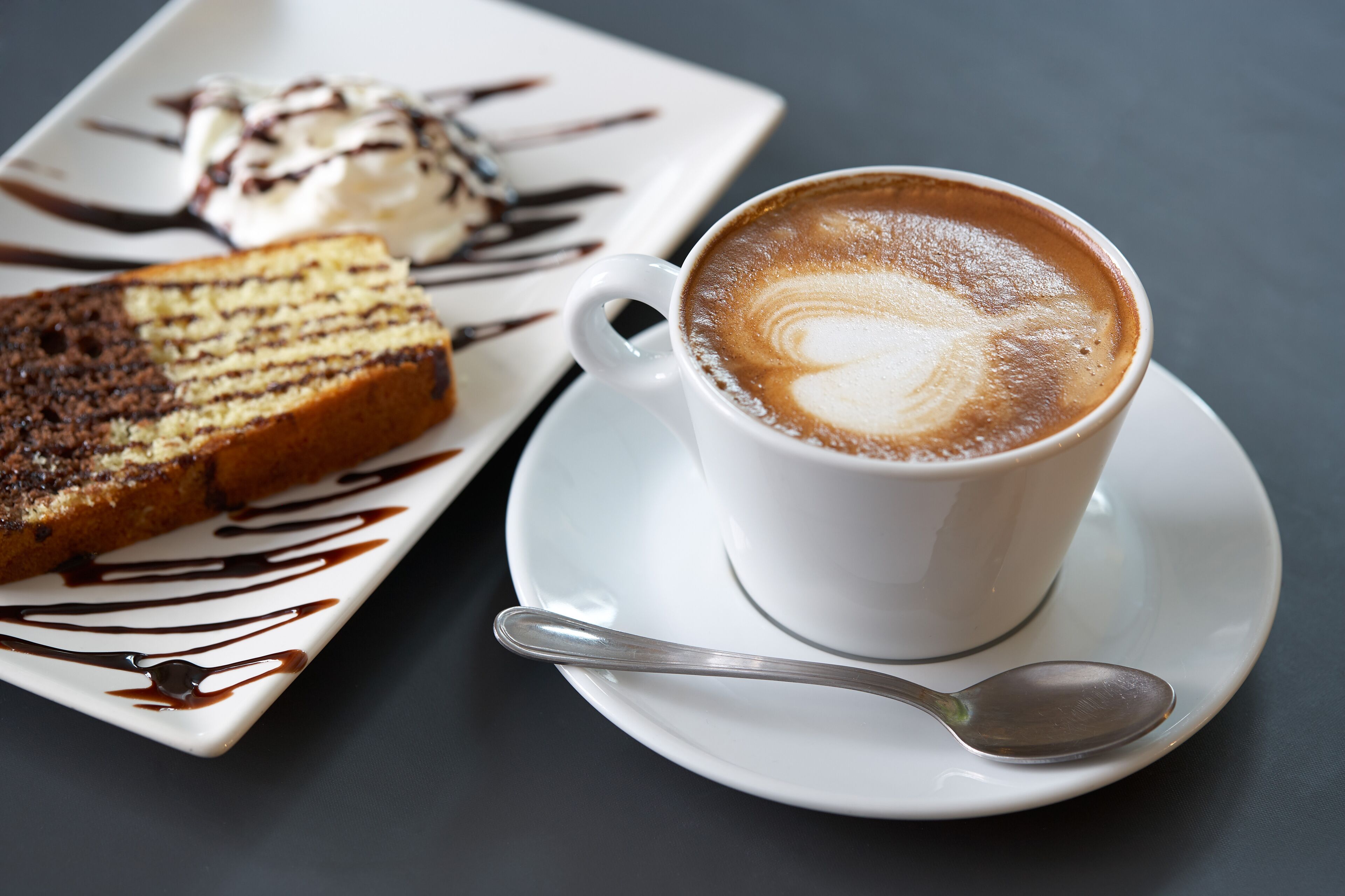 Una taza de café con arte de latte sobre un platito, una porción de pastel con glaseado de chocolate y una cuchara sobre un plato blanco.