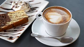 Una taza de café con arte de latte sobre un platito, una porción de pastel con glaseado de chocolate y una cuchara sobre un plato blanco.