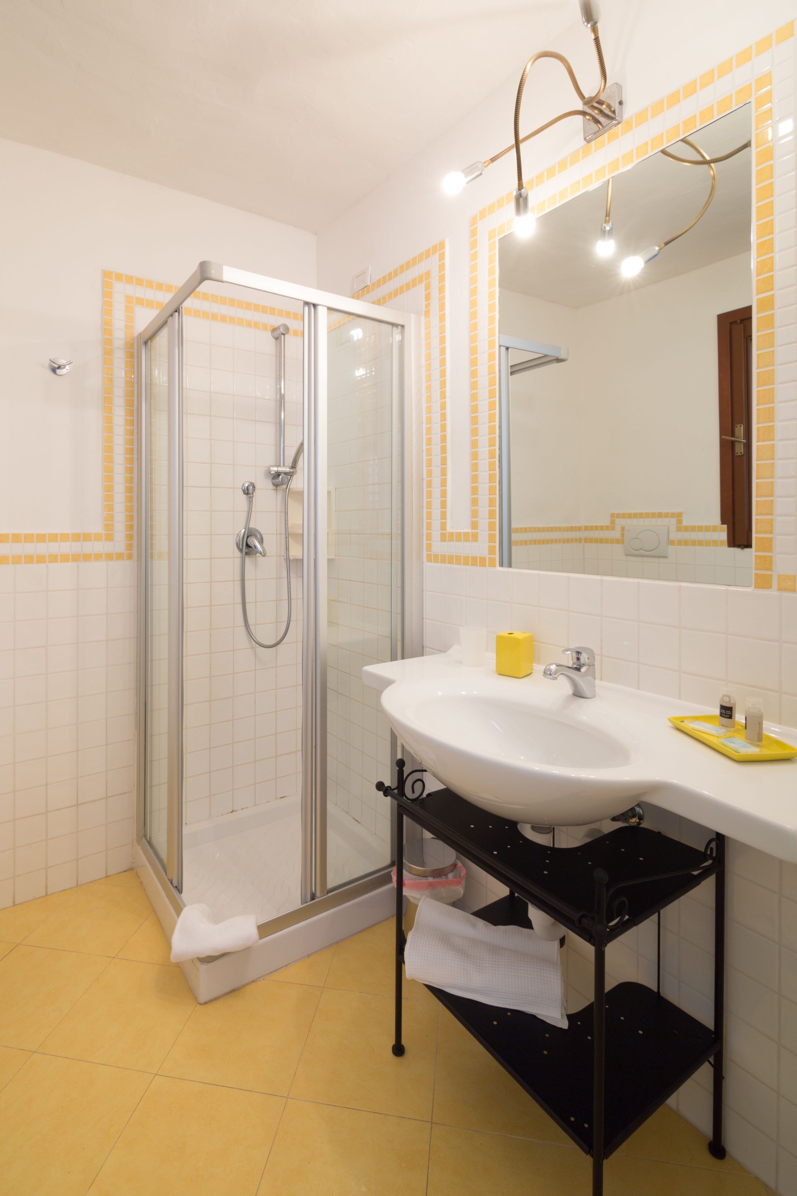A bathroom with a glass-enclosed shower, a white sink with a yellow soap dispenser, and a mirror with yellow-tiled borders.