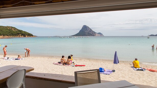 A beach view with a mountain in the distance, people sunbathing, and beach umbrellas. - White Sand Residence (Olbia)