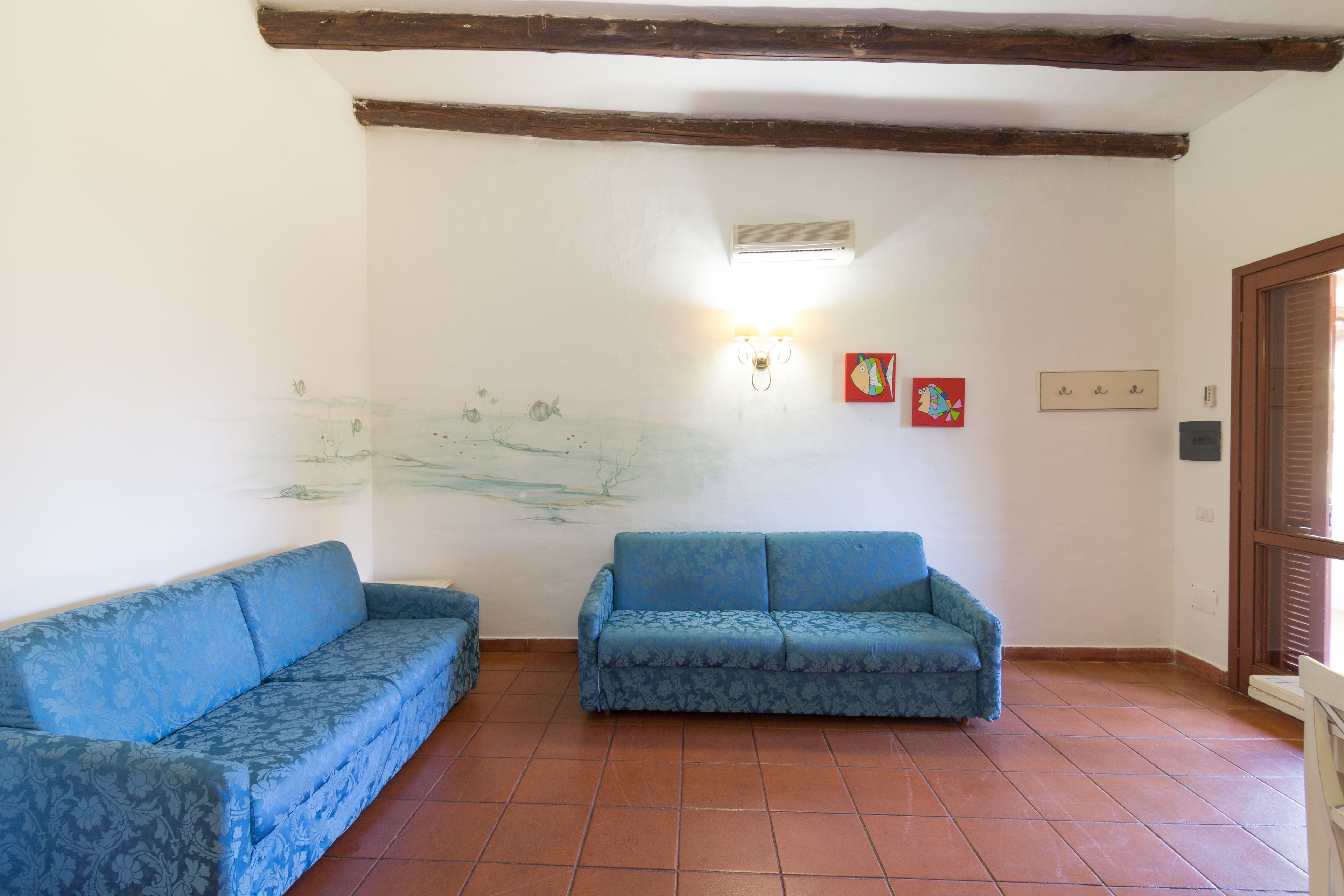 A room with two blue patterned sofas, a tiled floor, a white wall with a mural, and a wooden ceiling beam.