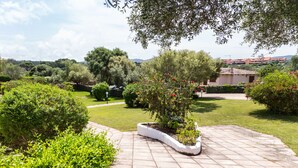 A paved garden area with a central planter, surrounded by lush greenery and a distant view of a building with red-tiled roofs. - White Sand Residence (Olbia)