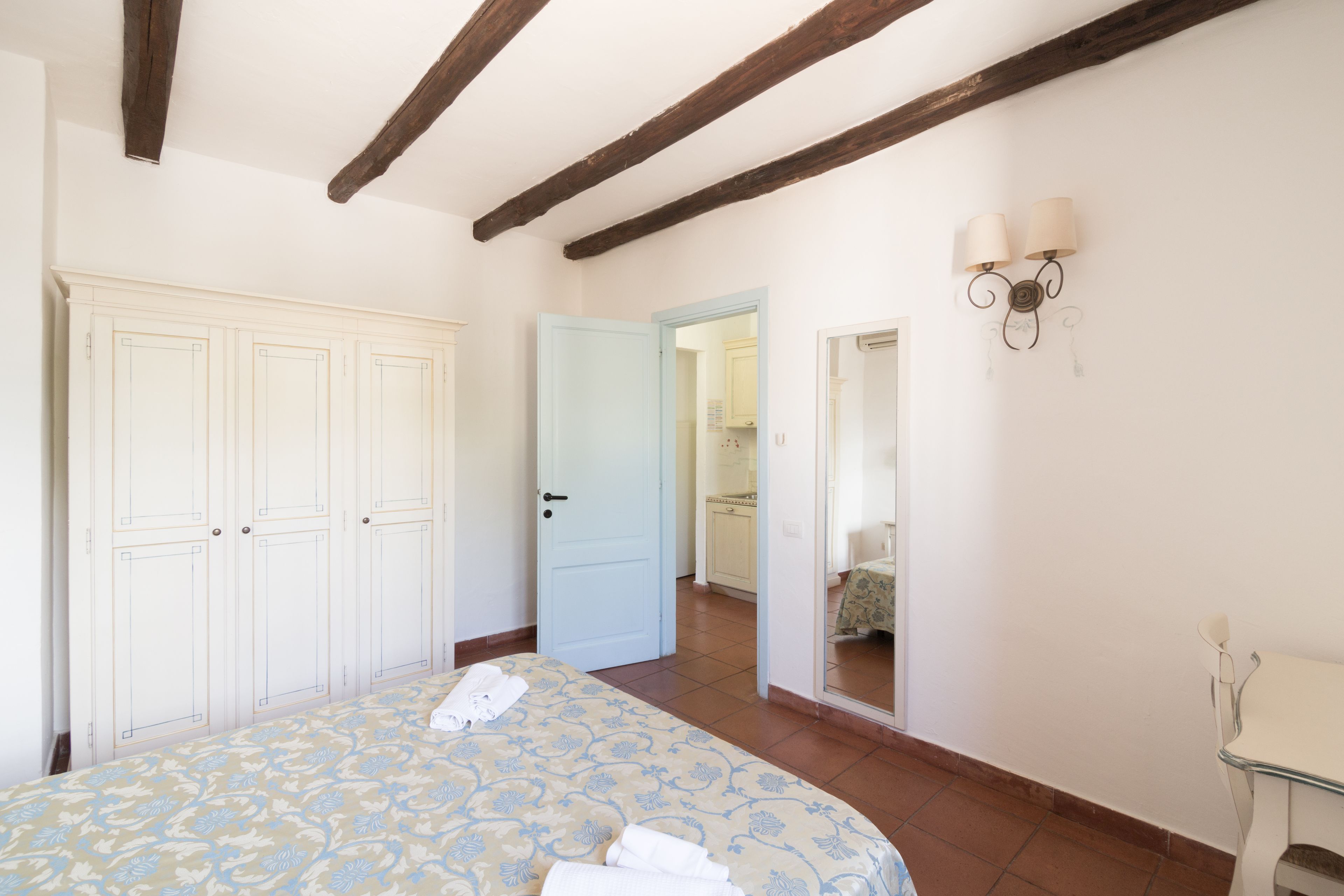 A bedroom with a bed, a wardrobe, a bathroom mirror, and a door leading to another room.