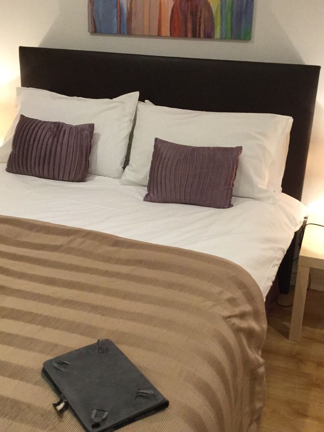A bed with white linens, two purple pillows, and a brown striped blanket. A black tablet is on the bed.