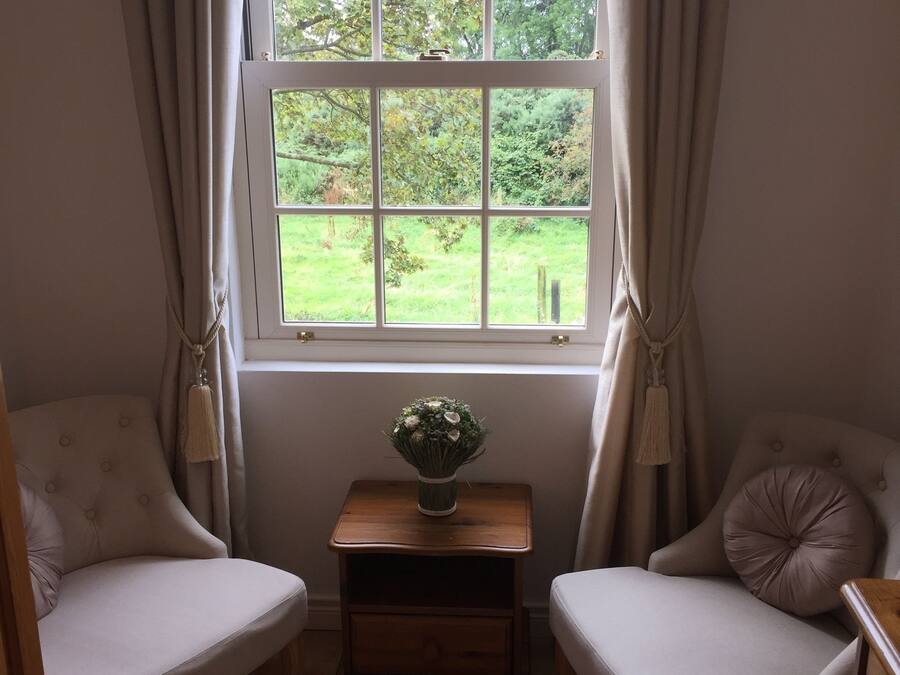 Una habitación con dos sillones, una pequeña mesa de madera con un jarrón y una ventana con cortinas.
