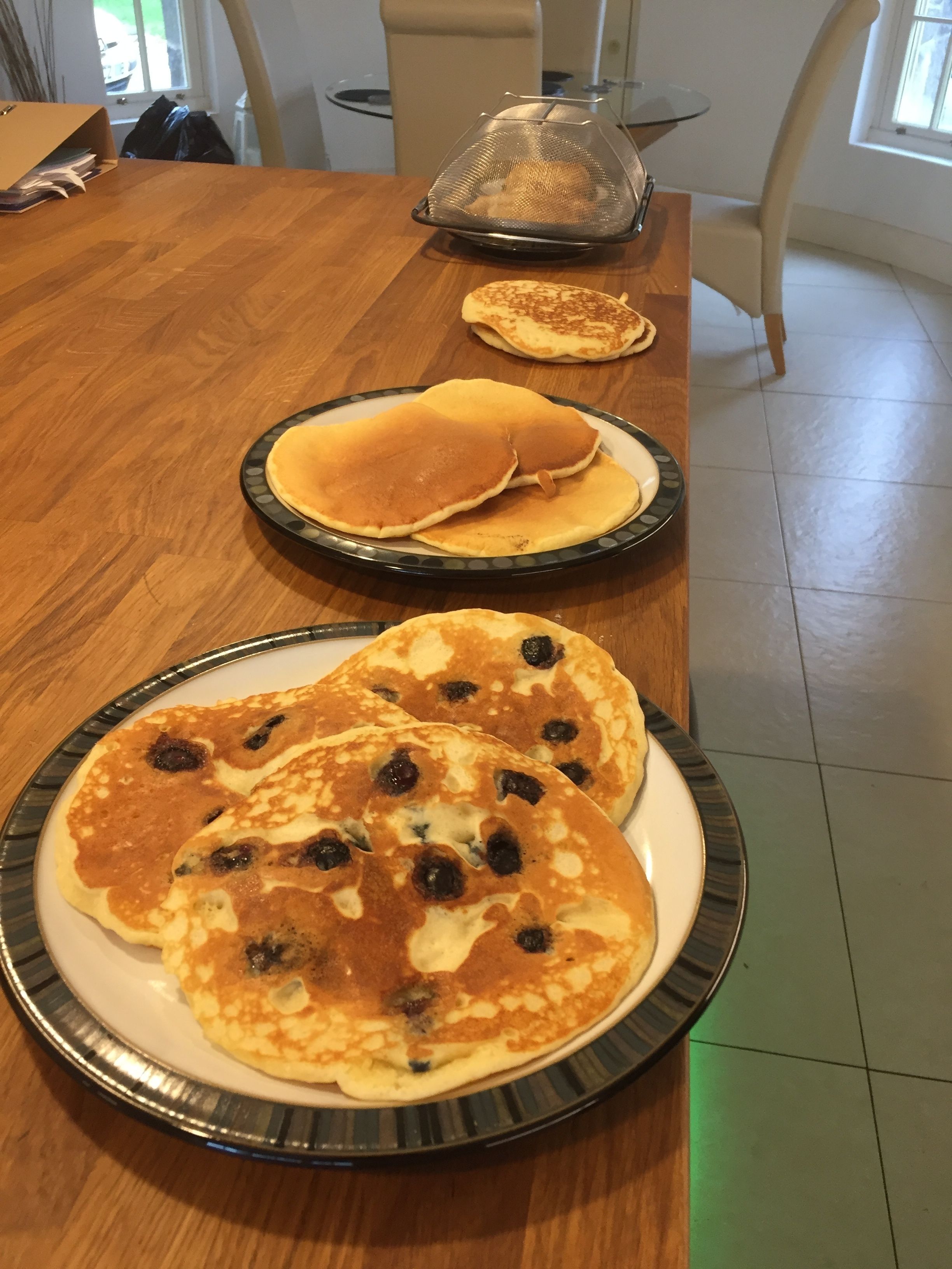 A dining table with three plates of pancakes, a colander, and a chair.