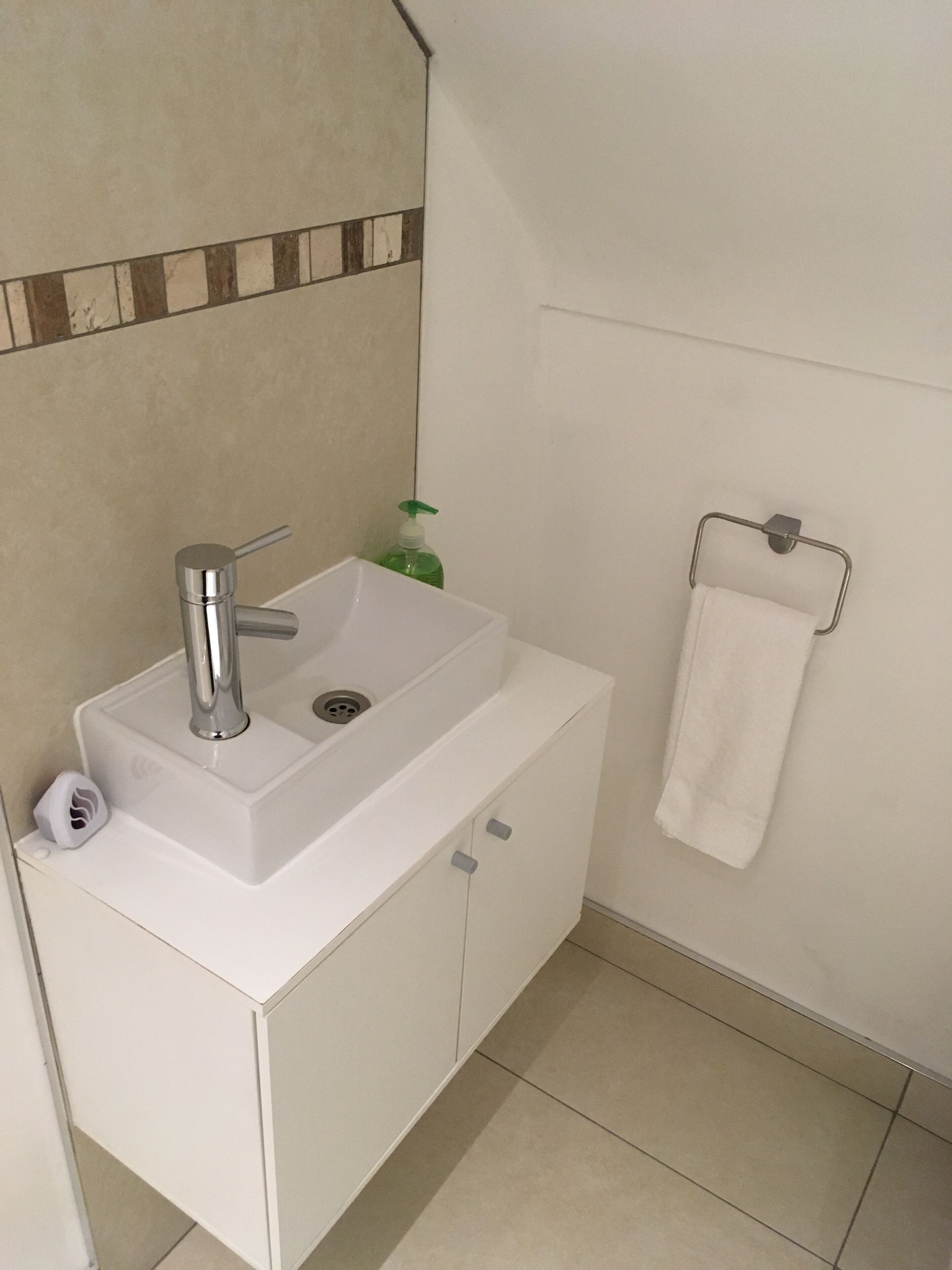 A bathroom with a white sink, a green soap dispenser, a white towel hanging on a metal rack, and a tiled floor.