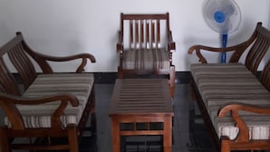 A room with a wooden sofa set, a matching armchair, and a small table. There is a blue fan on the floor. - Hotel Blue Moon (Dambulla)