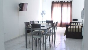 Een kamer met een eettafel en stoelen, een aan de muur bevestigde televisie, een ventilator en een raam met gordijnen.
