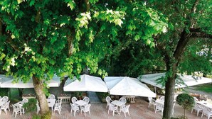 Une terrasse extérieure avec des chaises et des tables en plastique blanc, protégée par des parasols blancs, et entourée d’arbres.