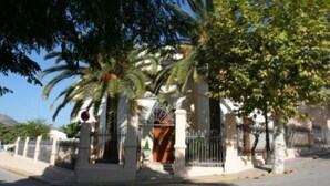 Una entrada protegida por un portón con una palmera y un edificio de fachada blanca.