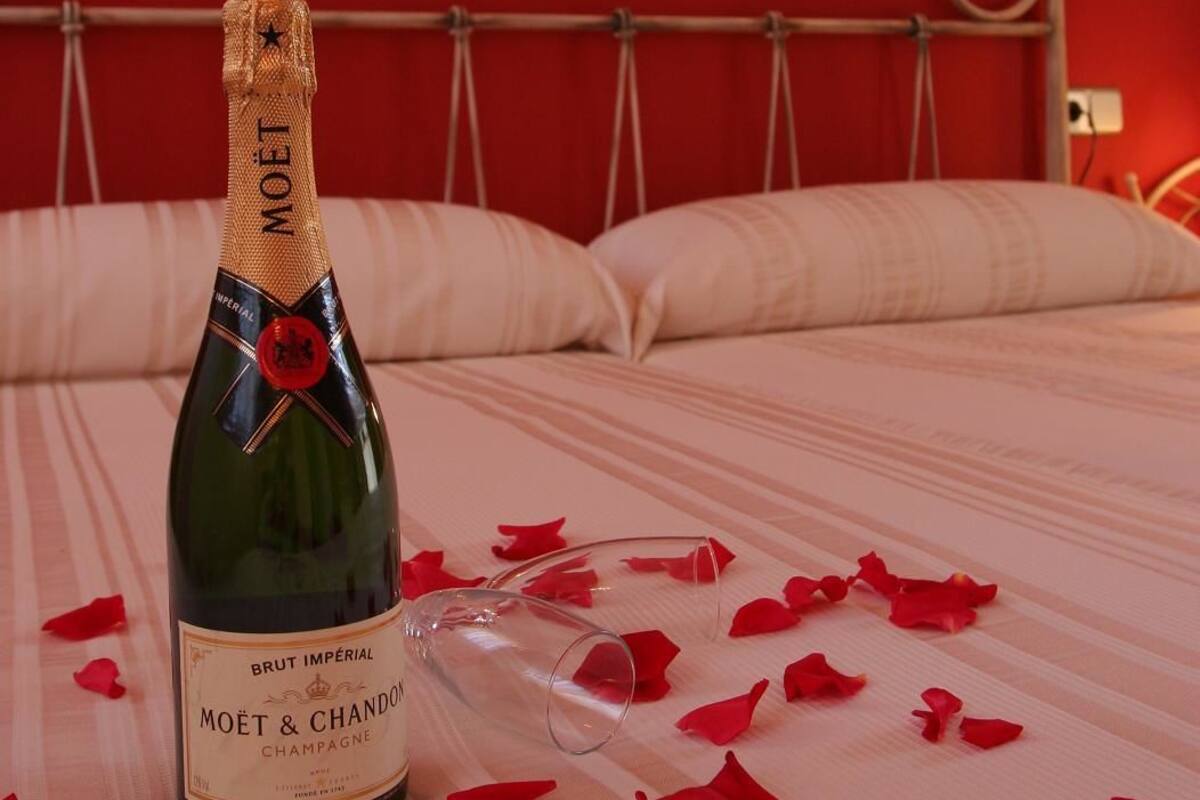 A bottle of Moët & Chandon champagne on a bed with rose petals, in a room with red walls and a metal bed frame.