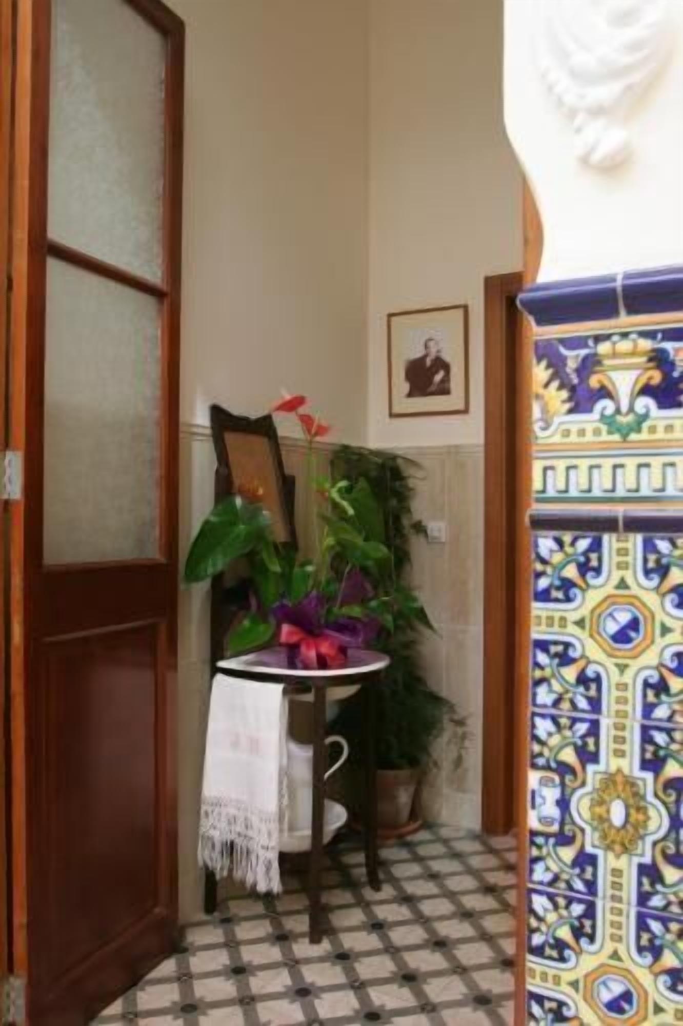 Un pasillo estrecho con suelo de baldosas, una puerta de madera, una pequeña mesa con mantel blanco, una planta en maceta y un cuadro colgado en la pared.