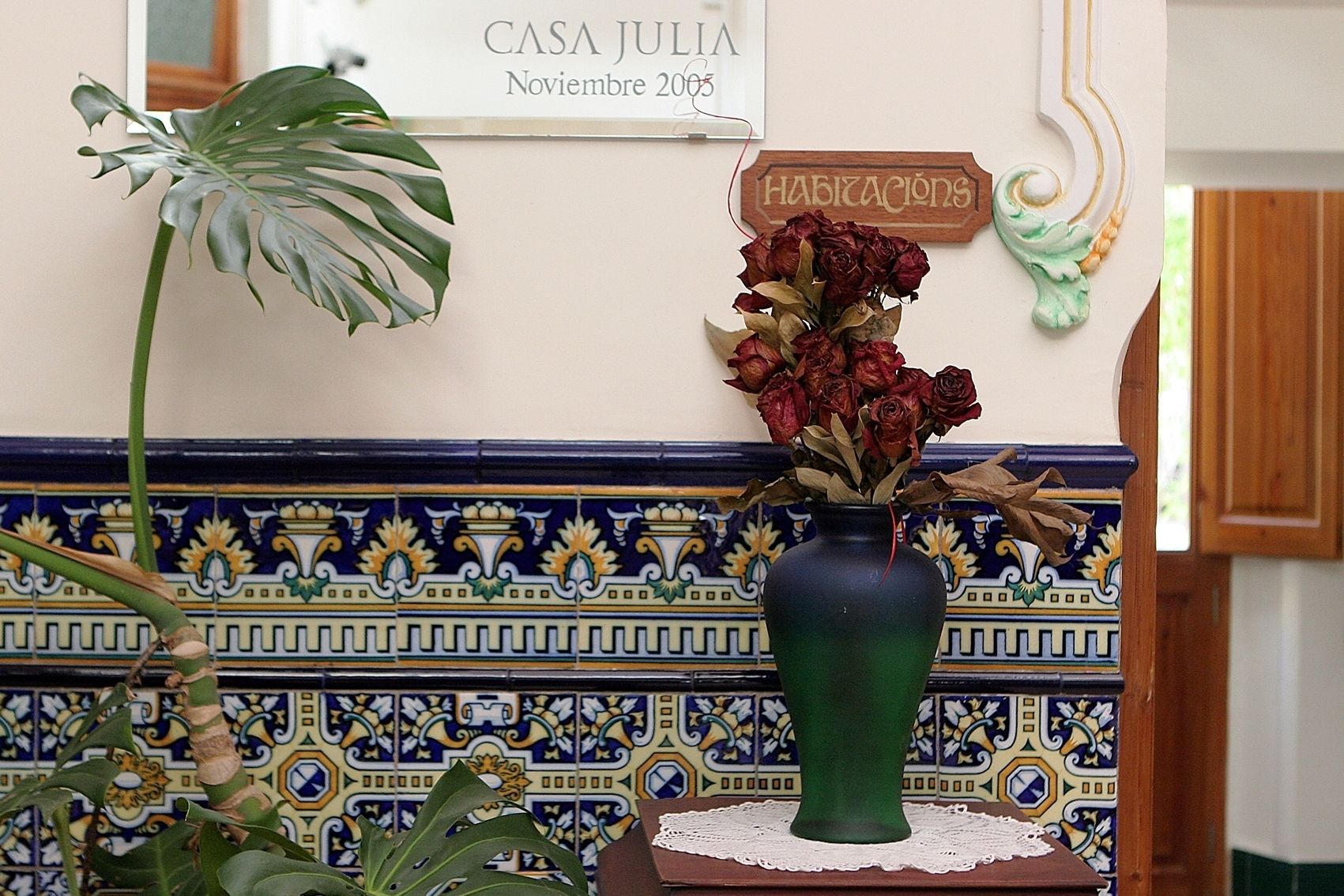 Un jarrón verde con rosas secas sobre una mesa, un letrero que dice "CASA JULIA" y una pared de azulejos decorativos.