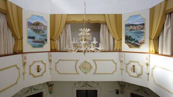 A grand room with ornate gold detailing, a chandelier, and two windows with curtains.