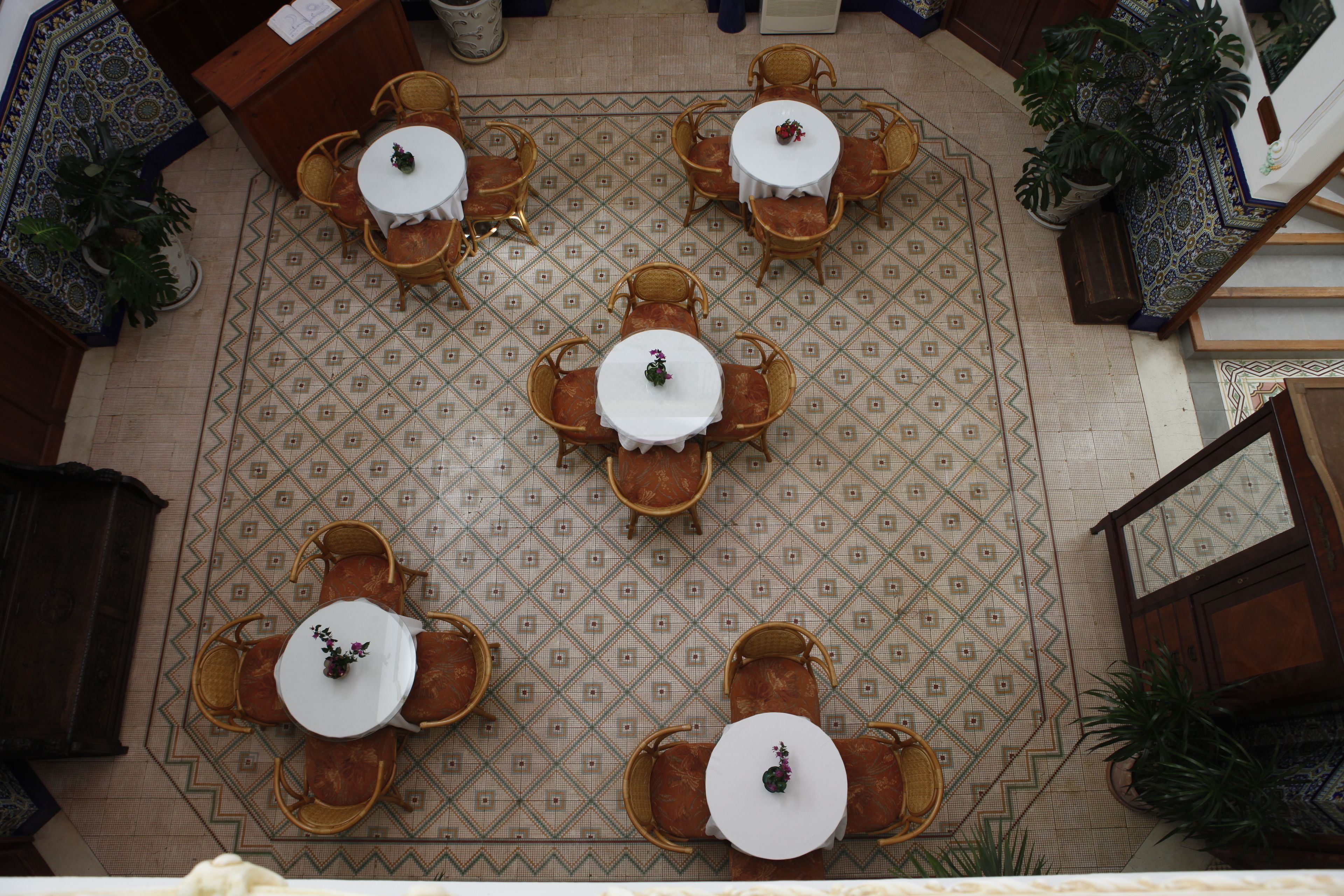 An overhead view of a hotel lobby with a patterned floor, multiple round tables with chairs, and potted plants.