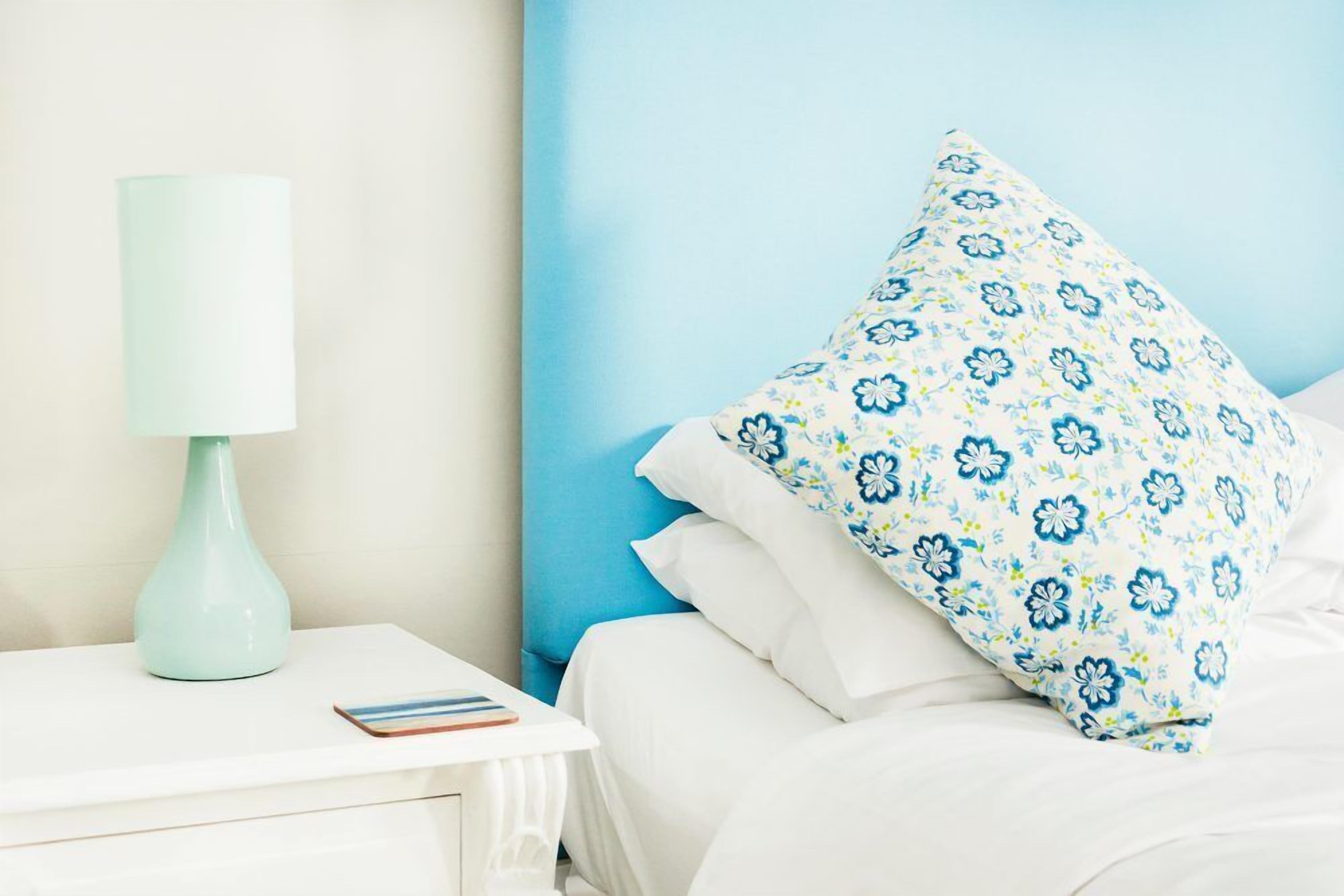 A neatly made bed with white bedding and a blue floral pillow, a white bedside table with a teal lamp, and a blue wall.