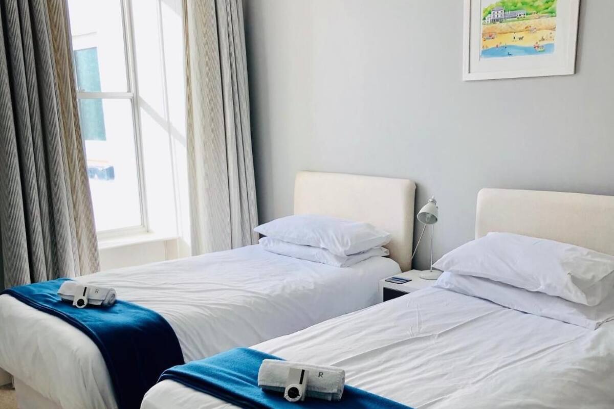 A hotel room with two beds, each with white linens and a blue blanket. There is a framed picture on the wall and a window with curtains.