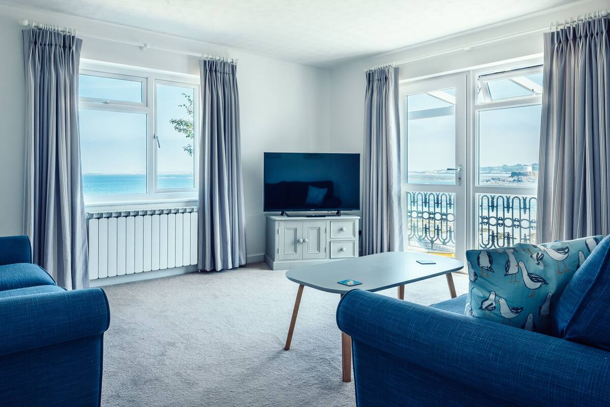 A modern living room with a large TV, two blue sofas, a white table, and a view of the sea through large windows.