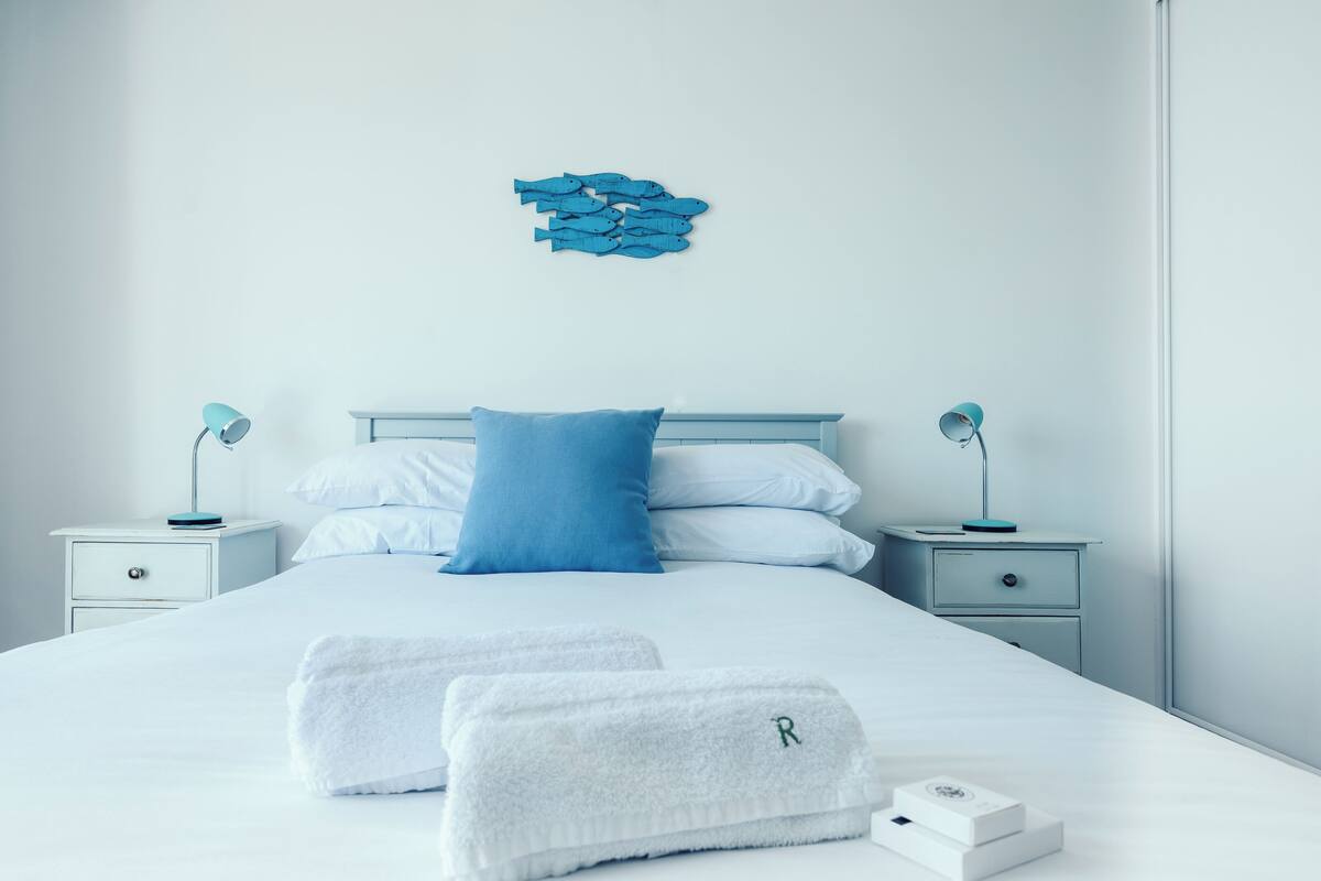 A neatly made bed with white linens and a blue pillow, two bedside tables with lamps, a wall art piece, and two towels on the bed.