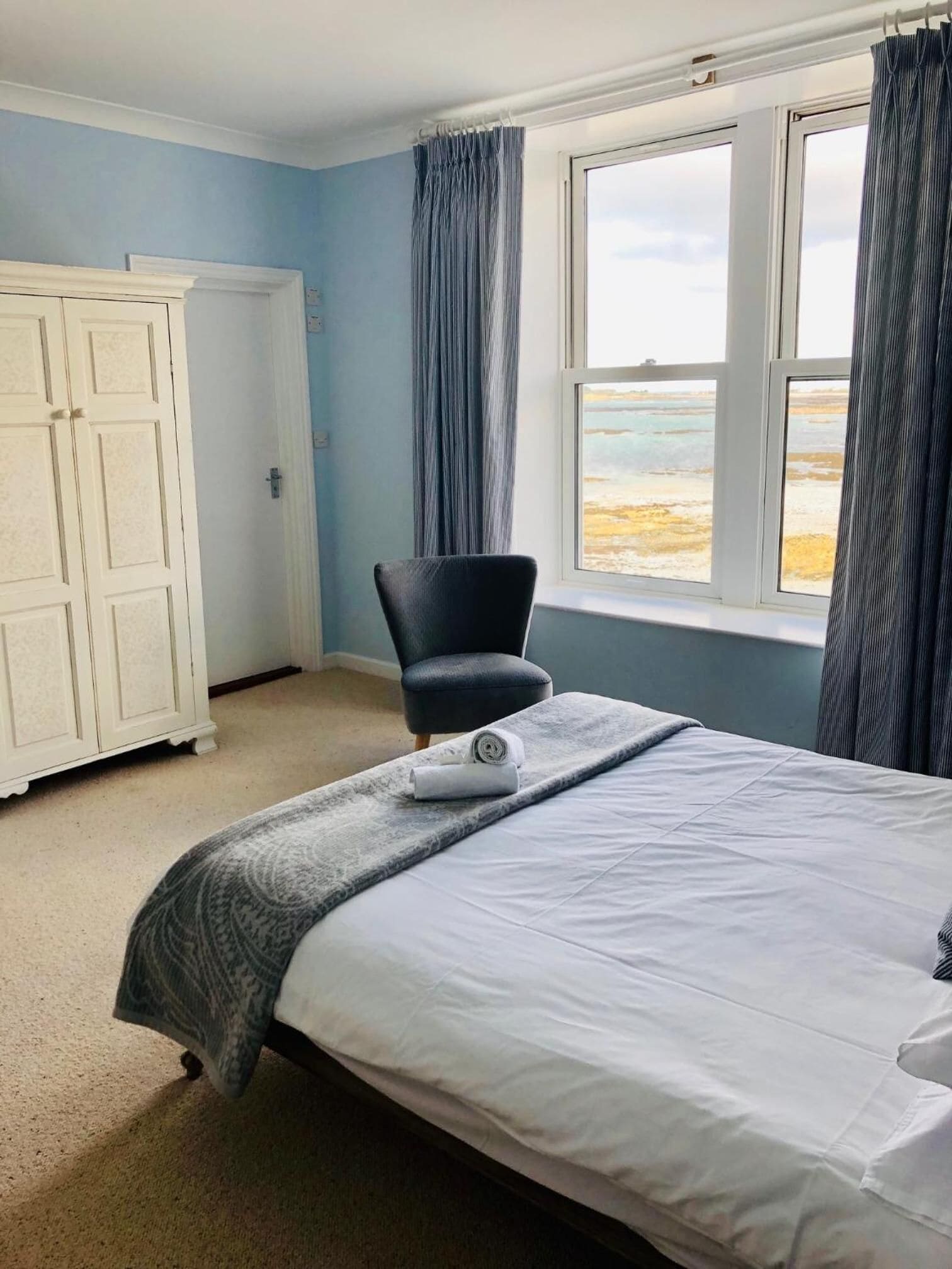 Een keurig opgemaakt bed met een opgerolde handdoek, een stoel en een garderobe in een kamer met een groot raam en uitzicht op het strand.