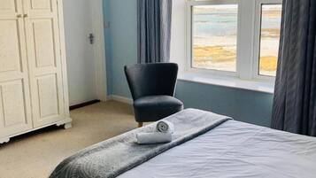 Een keurig opgemaakt bed met een opgerolde handdoek, een stoel en een garderobe in een kamer met een groot raam en uitzicht op het strand.