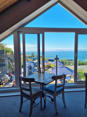A dining area with a table and chairs, large windows with a view of the sea and a coastal town. - The Imperial Hotel (Torteval)