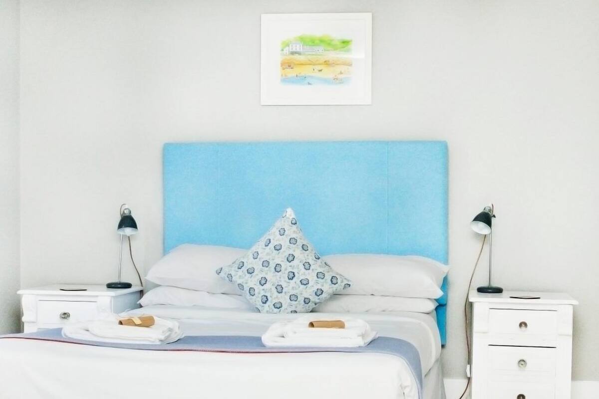 A neatly made bed with a blue headboard, white pillows, and a single patterned pillow. There are two bedside tables with lamps, a white nightstand, and a framed picture on the wall.