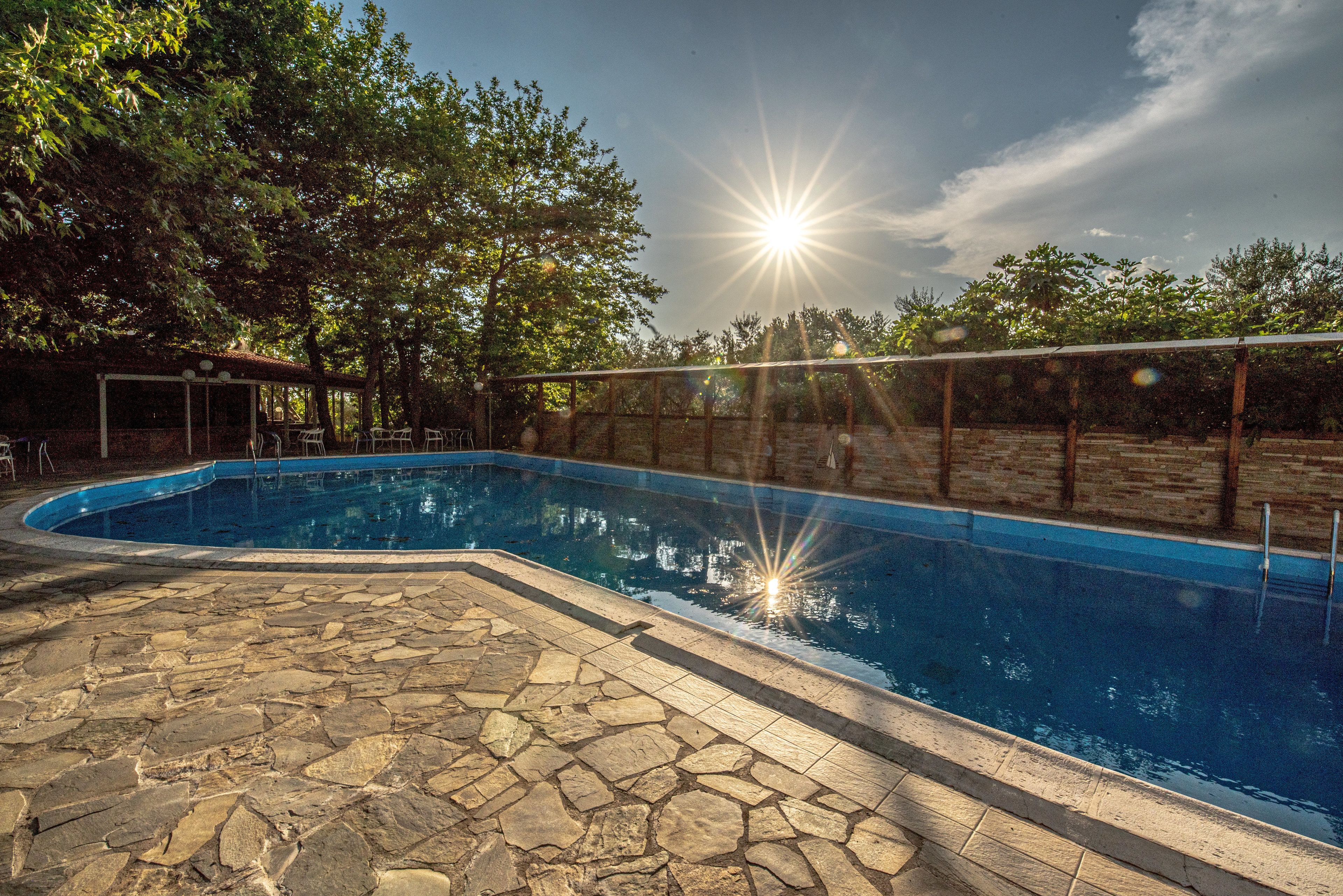 A swimming pool with stone tiles, surrounded by trees and a covered seating area.