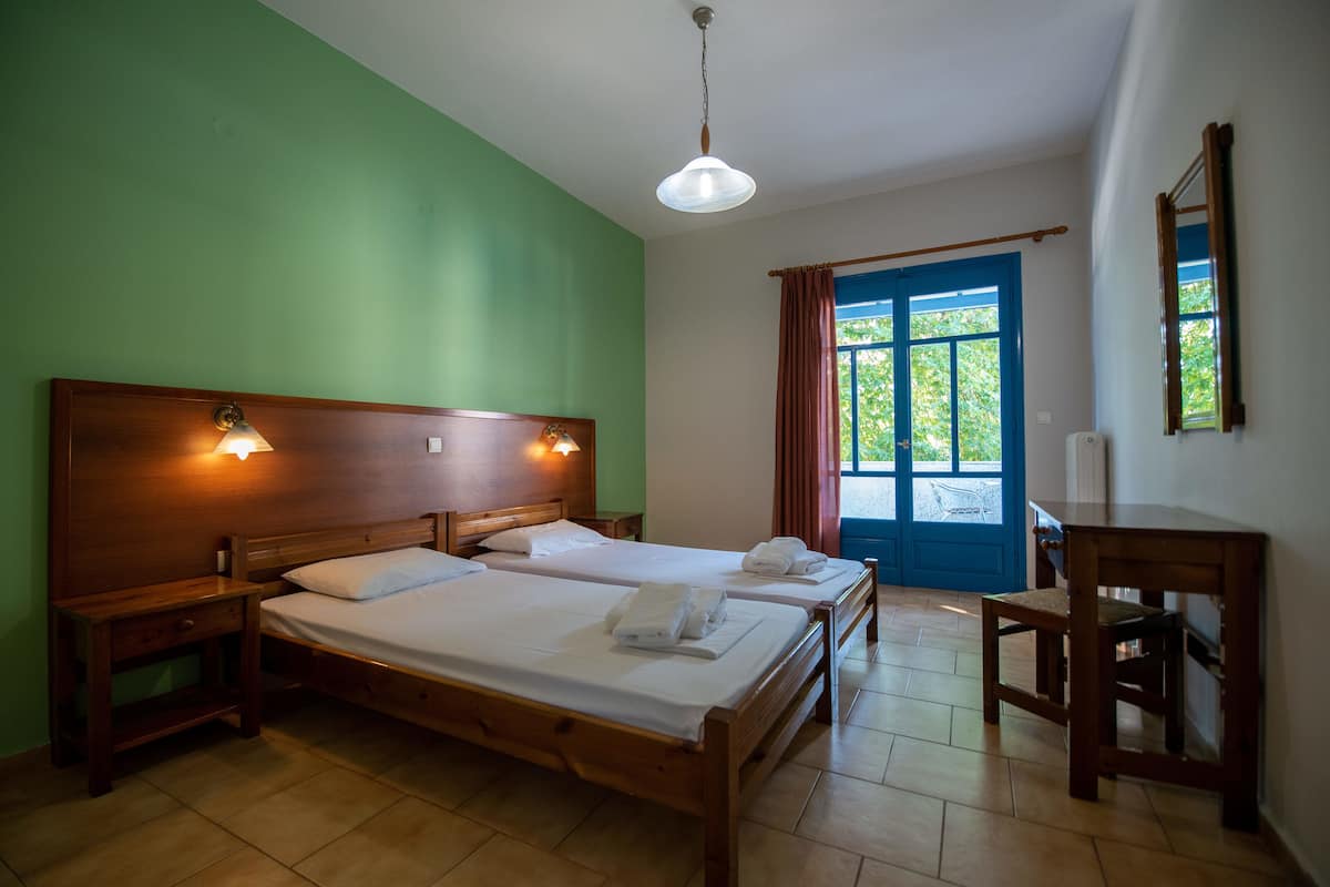 Una habitación con dos camas de madera, una mesita de noche de madera, una silla de madera, una ventana con cortinas azules y una lámpara en el techo.