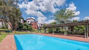 A swimming pool with a wooden pergola, surrounded by a brick pathway and greenery. - Jackal Creek Self-Catering Apartments (Roodepoort)