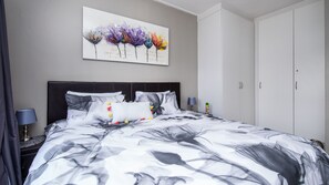 A bedroom with a bed, a nightstand, a lamp, a wardrobe, and a painting on the wall. - Jackal Creek Self-Catering Apartments (Roodepoort)