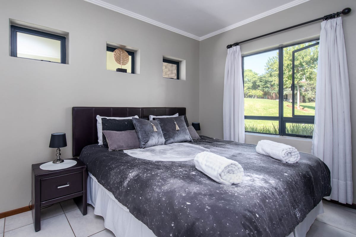 A bedroom with a large bed, a nightstand, a window with curtains, and a view of greenery outside.