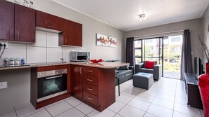 A modern kitchen with wooden cabinets, a microwave, and a built-in oven. There is a dining area with a grey sofa and a red cushion. - Jackal Creek Self-Catering Apartments (Roodepoort)