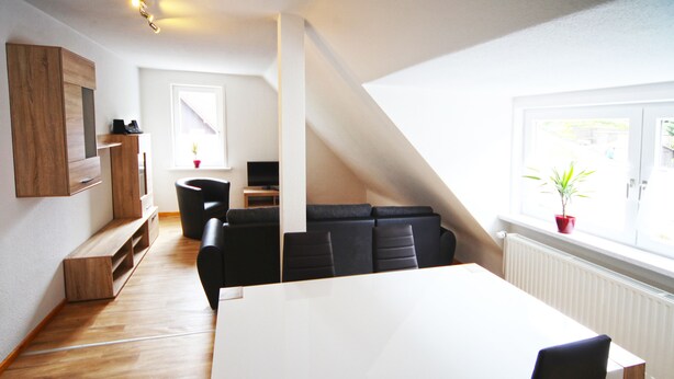 A modern living room with a large white table, black chairs, and a wooden bench. There is a TV and a plant on a shelf. - FeWo Lerbachtal (Osterode am Harz)