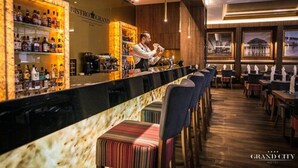 A bar area with a bartender, shelves of liquor bottles, and a view of a building through large windows. - Grand City Hotel Wrocław (Wroclaw)