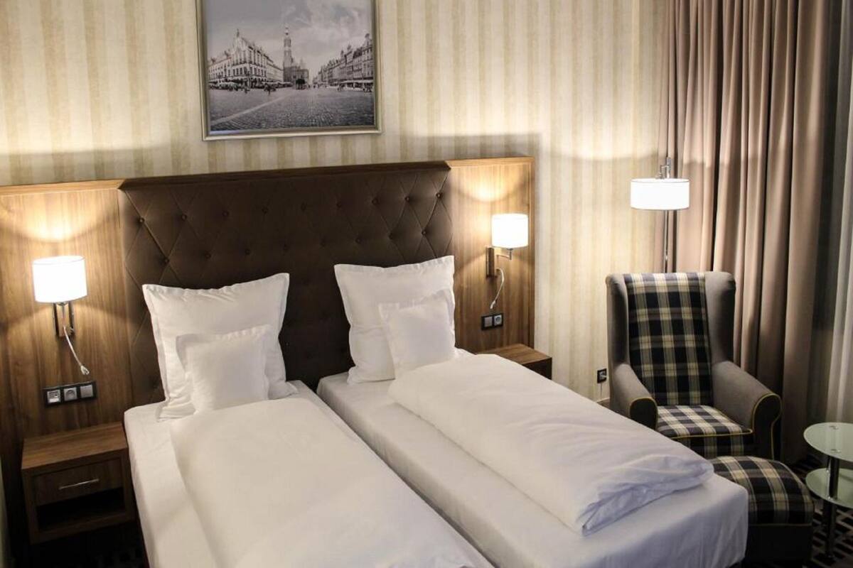 A hotel room with two beds, a headboard, nightstands, a chair, a small table, and a wall-mounted picture.