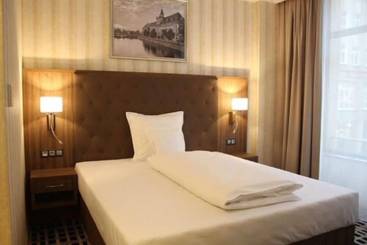 A hotel room with a large bed, two bedside lamps, a headboard, a nightstand, and a window with curtains.