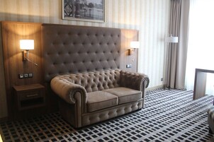 Habitación de hotel con cabecera tapizada, sofá, dos lámparas de noche, mesitas de noche pequeñas y suelo estampado.