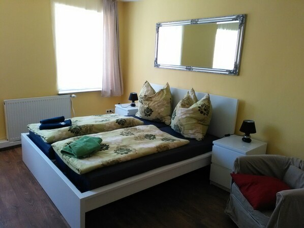A double bed with floral bedding, two bedside tables with lamps, a mirror above the bed, a radiator, and a window with curtains.