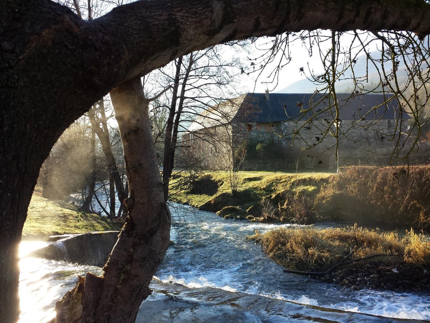 A river flowing through a rural landscape with a stone bridge, a stone house, and trees framing the scene.