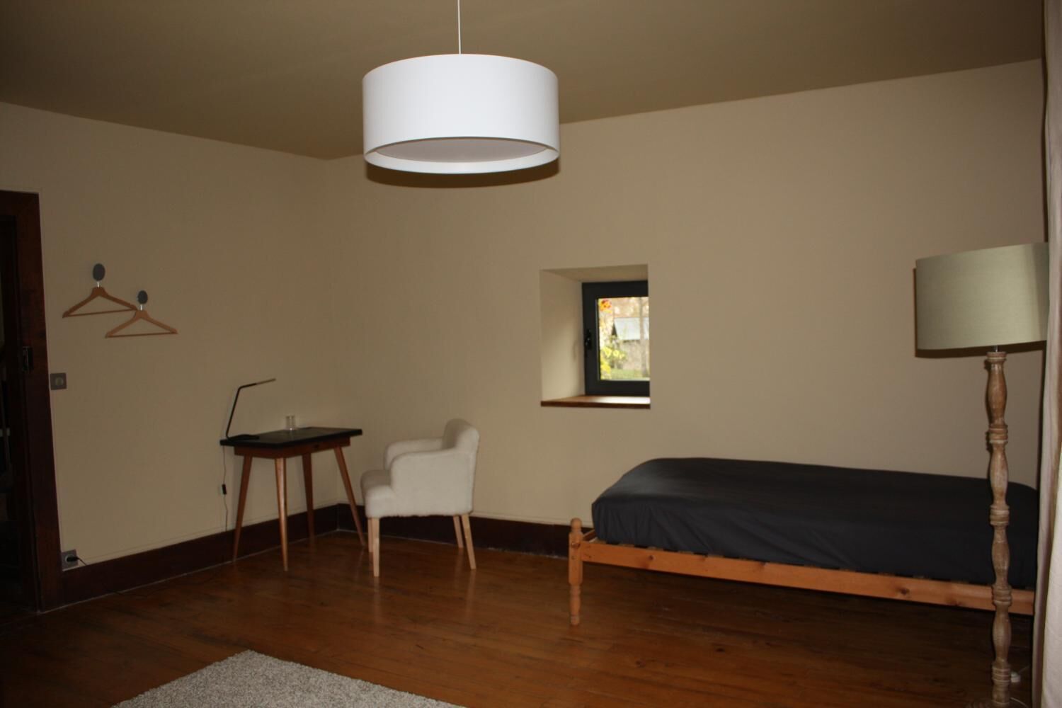 Un dormitorio con cama, escritorio, silla, lámpara colgante, ventana y piso de madera.