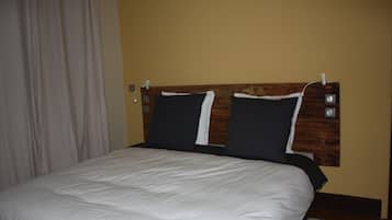 Un letto rifatto con cura, una testiera in legno, un muro semplice e una tenda a sinistra.