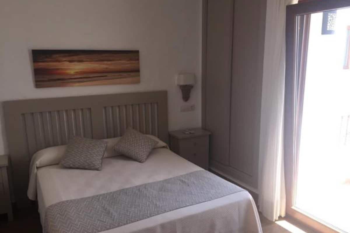 Quarto com cama, mesa de cabeceira, luminária na parede e janela com cortinas.